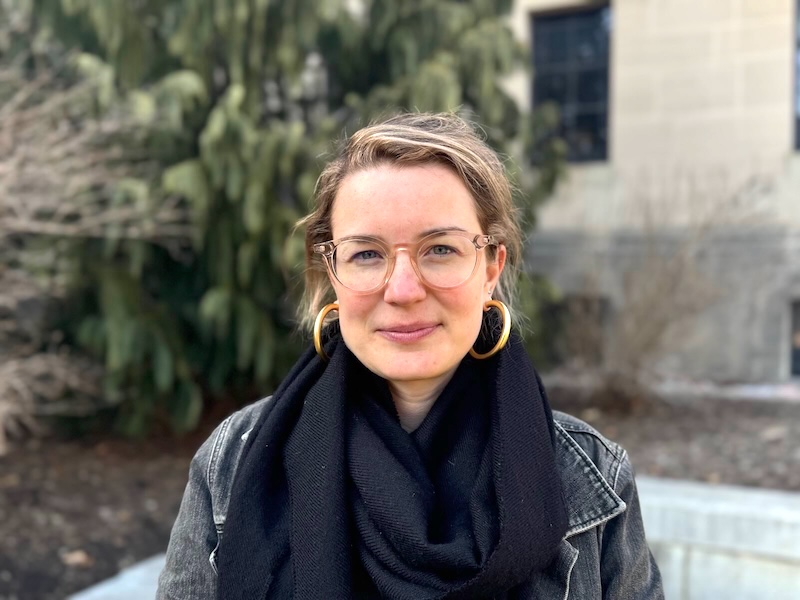 A portrait of a 30-something white woman with glasses and large gold hoop earrings looks into the camera and smiles slightly. The photo is outside, with a brick building and a tree in the background.